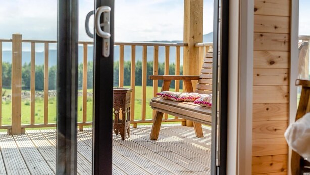 Eine Holzterrasse mit einer Bank und einem kleinen Tisch, Blick auf eine grüne Landschaft und eine Holzwand mit Lichtschalter.