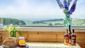 Ein Tisch mit Brot, Honig und Spirituosenflaschen, ein Honigglas und eine Vase mit Blumen an einem Fenster mit Blick auf eine Landschaft.
