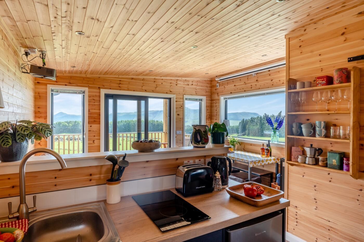 A wooden cabin kitchen with a large window, a sink, a stove, and a shelf with kitchenware.