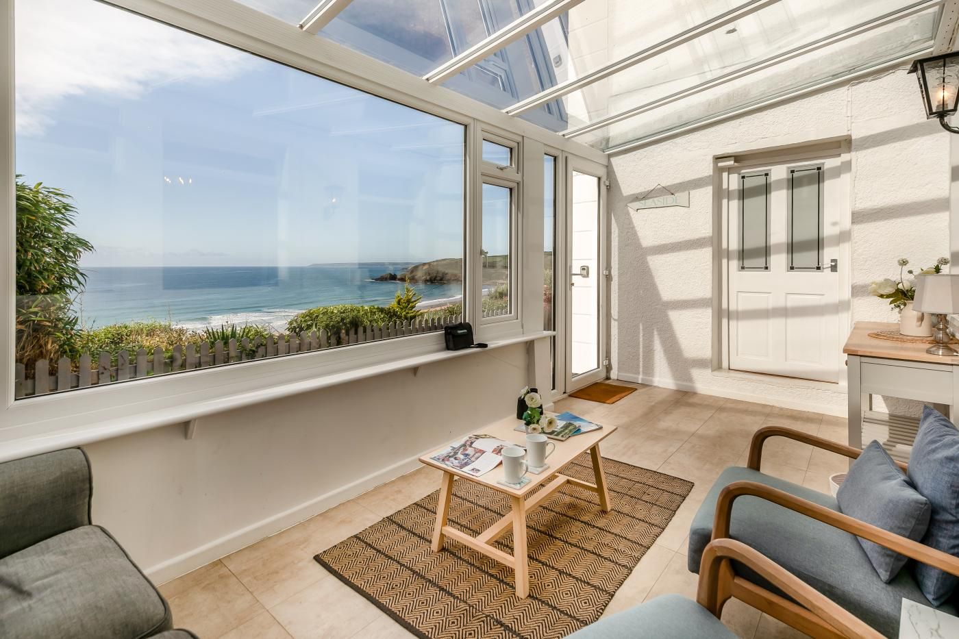 A room with a large window overlooking the sea, a small table with a rug, and a door leading to another room.