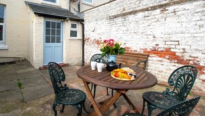 Een terras met een ronde houten tafel, twee stoelen, een blauwe deur en een bakstenen muur.