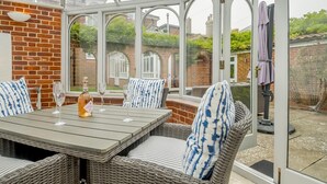 A conservatory with a brick wall, wicker furniture, and a table with wine glasses and a bottle. - 5 Bed in Southwold (oc-tomill) (Southwold)