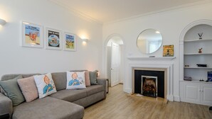 A living room with a grey sectional sofa, a fireplace, and a white wall with framed posters. - 5 Bed in Southwold (oc-tomill) (Southwold)