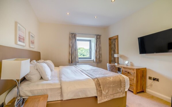 A bedroom with a bed, bedside lamp, wooden dresser, TV, and a window with curtains.