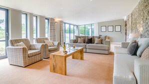 A spacious living room with large windows, a stone wall, and a mix of sofas and chairs. - 4 Bed in Catsash (oc-u32332) (Newport)
