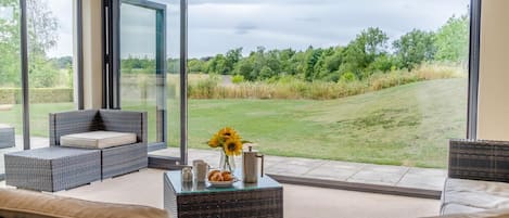 A modern living room with wicker furniture, a glass coffee table, and a view of a grassy landscape through large glass doors.