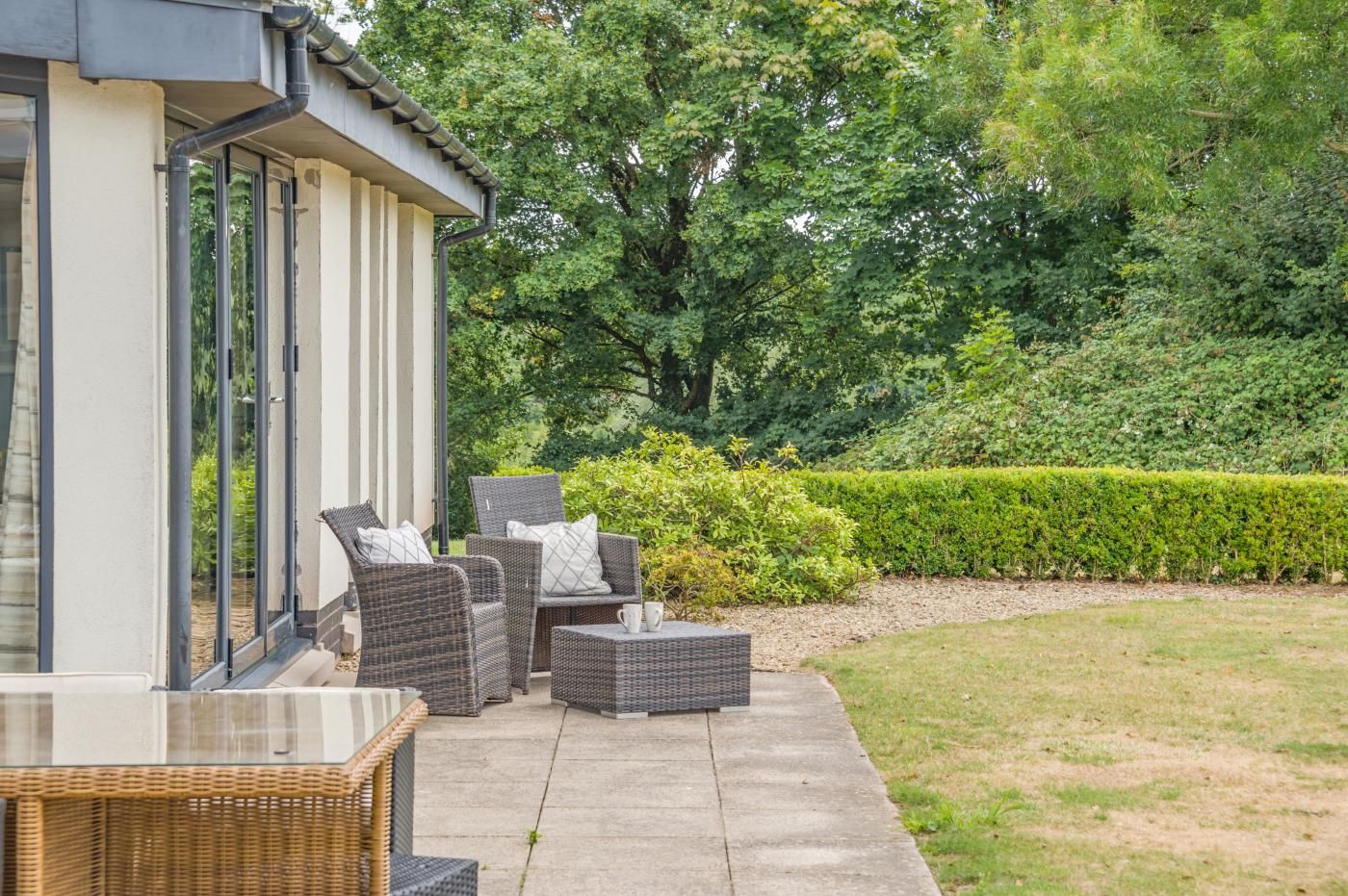 A patio with wicker furniture, a glass table, and a hedge in the background.