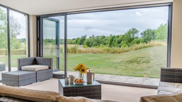 A modern living room with wicker furniture, a glass coffee table, and a view of a grassy landscape through large glass doors. - 4 Bed in Catsash (oc-u32332) (Newport)