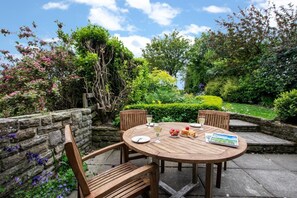 Eine Terrasse im Freien mit einem runden Holztisch und vier Holzstühlen, umgeben von üppiger Bepflanzung und blühenden Pflanzen.