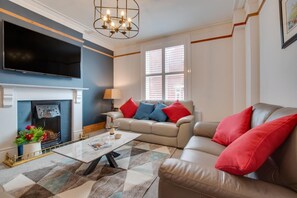 Ein Wohnzimmer mit Kamin, einer Couch mit roten und blauen Kissen, einem Couchtisch und einem an der Wand montierten Fernseher.