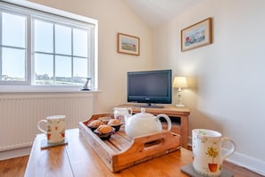 A cozy room with a large window, a flat-screen TV, a wooden side table with a tray of muffins, a teapot, and two fruit-themed mugs. - 2 Bed in Whitby (oc-w010) (Whitby)