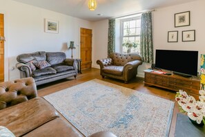 A living room with a sofa, armchair, and ottoman, a television, and a large window with curtains. - 2 Bed in Higher Penponds (oc-h32383) (Camborne)