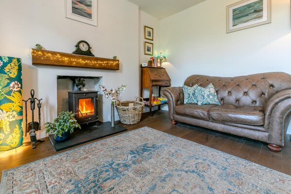 A cozy living room with a fireplace, a brown leather sofa, a patterned rug, and a wooden shelf with lights. - 2 Bed in Higher Penponds (oc-h32383) (Camborne)