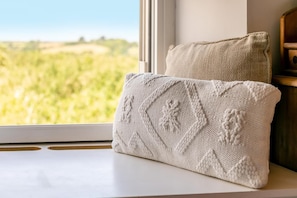 Ein Fensterplatz mit Blick auf ein grünes Feld, ein weißes Strickkissen und ein Holznachttisch.