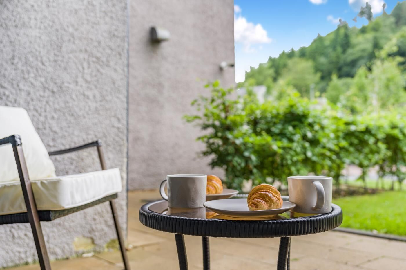 A round table with two white mugs and a croissant on a plate, set against a backdrop of a stone wall and greenery.