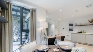 Un comedor moderno con una mesa dispuesta para dos personas, una ventana con cortinas, una cama con cabecera y una cocina con fregadero y armarios.