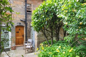 A stone building with a wooden door, a small outdoor seating area with chairs, and a garden with yellow flowers. - 2 Bed in Whitby (oc-r038) (Robin Hood's Bay)