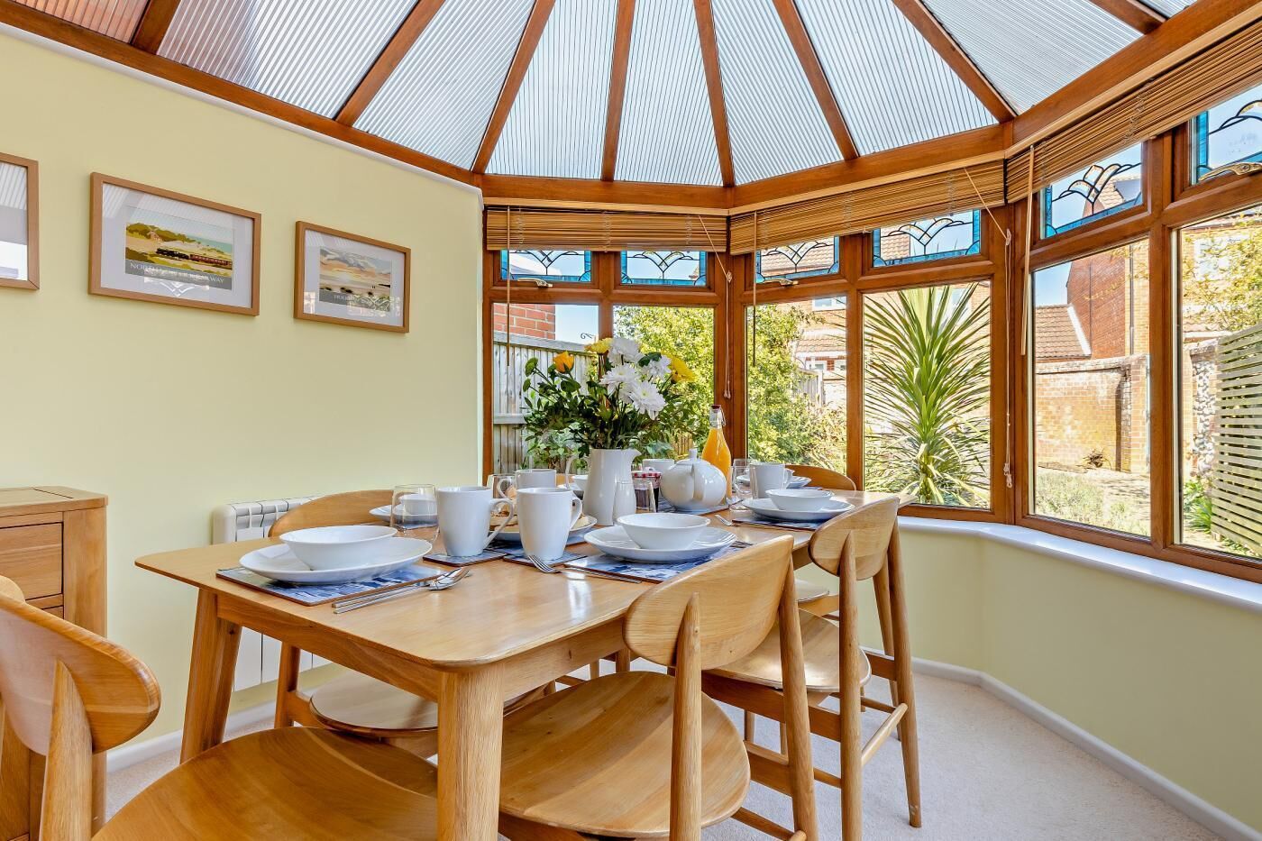 A dining area with a wooden table set for four, surrounded by chairs. There are framed pictures on the wall, a window with a view of greenery, and a glass ceiling structure.