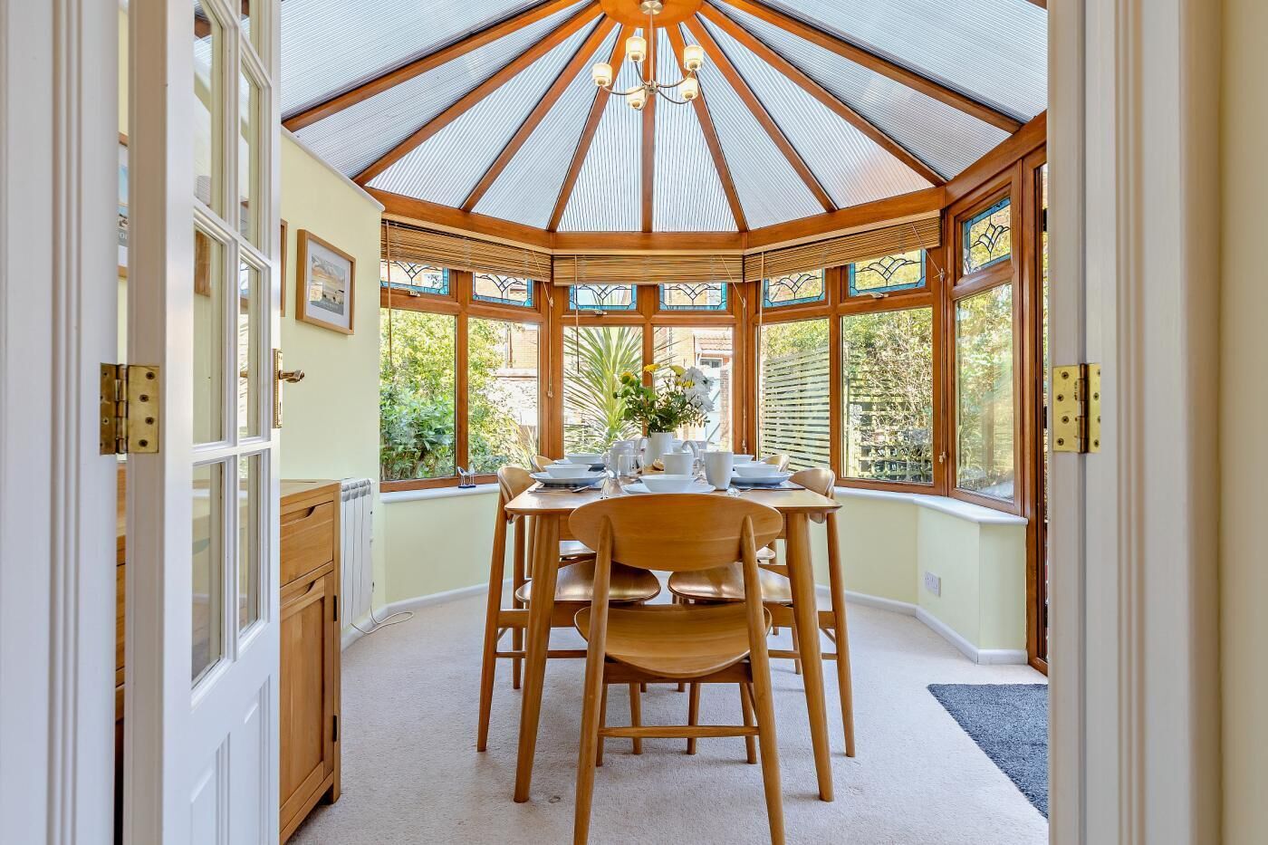 A dining area with a wooden table and chairs, a glass ceiling, and large windows offering a view of the outdoors.