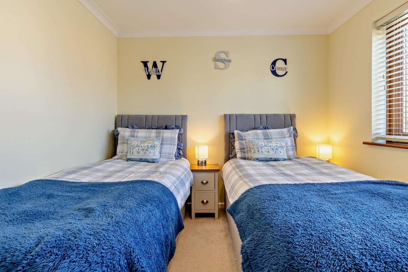 A bedroom with two beds, each with a blue blanket, a bedside table with a lamp, and wall decorations with letters W, S, and C.