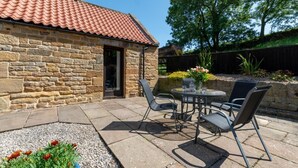A stone building with a tiled roof, a patio with a round table and chairs, and a garden with flowers and a tree. - 2 Bed in Whitby (oc-y003) (England)