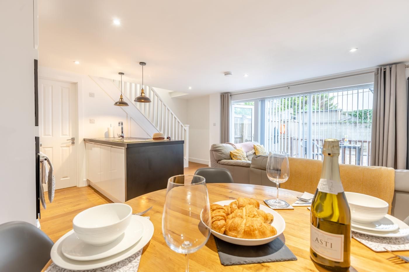 A dining area with a wooden table set for two, a bottle of wine, and a bowl of croissants. A kitchen is visible in the background with a white countertop and hanging pendant lights.