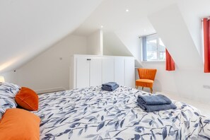 Ein Schlafzimmer mit schräg versiegelter Decke, einem Bett mit floralem Überwurf, einem orangefarbenen Sessel und einem Fenster mit roten Vorhängen.