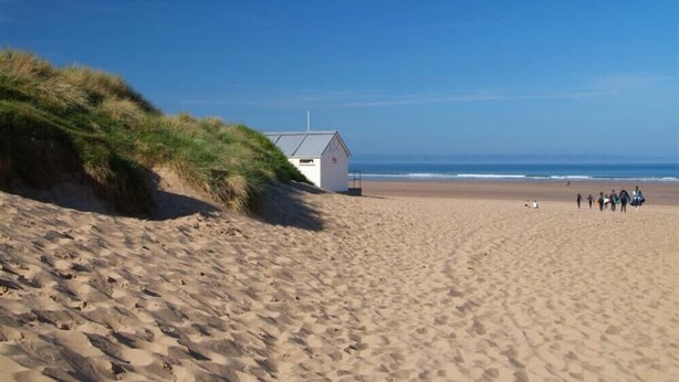 Ein Sandstrand mit einem kleinen weißen Gebäude, vermutlich eine Rettungsstation, und einem grasbewachsenen Dünenrücken.