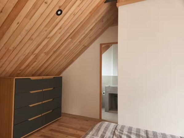 Un dormitorio con techo inclinado, un armario, una cama y un baño visible a través de una puerta abierta.