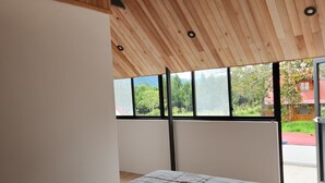 A bedroom with a wooden ceiling, a bed with a striped blanket, and a view of greenery outside through large windows.