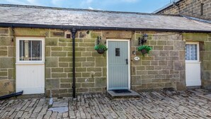 A stone building with a grey door, two windows, and a small porch area. - 2 Bed in Pateley Bridge (oc-ds127) (Castlestead)