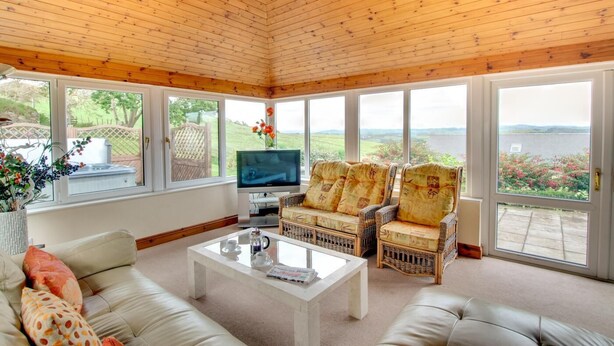 A spacious living room with a wooden ceiling, large windows, a flat-screen TV, wicker furniture, and a white coffee table. - 5 Bed in Aberystwyth (oc-wan443) (Llanfarian)