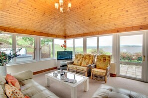 A spacious living room with a wooden ceiling, large windows, a flat-screen TV, wicker furniture, and a white coffee table. - 5 Bed in Aberystwyth (oc-wan443) (Llanfarian)