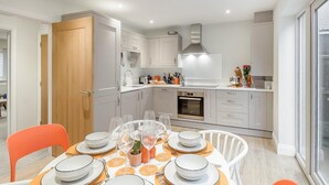 Una cocina moderna con armarios blancos, horno empotrado y zona de comedor para cuatro con sillas naranjas y una mesa dispuesta con platos, vasos y cubiertos.