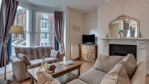 A living room with a sofa set, a coffee table with a bottle and flowers, a television on a wooden cabinet, and a fireplace with a mirror above it. - 2 Bed in Whitby (oc-w945) (Whitby)