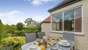 Een buitentafel gedekt met borden, kommen, glaasjes sinaasappelsap en een pot ontbijtgranen.