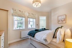 Ein Schlafzimmer mit einem Bett, zwei Fenstern mit blumigen Vorhängen, einem Heizkörper, einer Tür und einem gerahmten Bild an der Wand.
