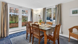A dining area with a table set for four, wooden chairs, a blue mat, and a view of a brick wall and greenery through glass doors. - 2 Bed in Whitby (oc-w793) (Whitby)