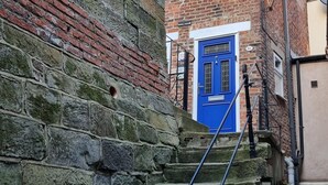 A narrow stone staircase leading to a blue door, with a brick wall on one side and a building on the other. - 2 Bed in Whitby (oc-w793) (Whitby)