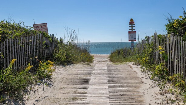 Ein hölzerner Steg führt zu einem Strand, im Hintergrund ist ein Leuchtturm zu sehen.