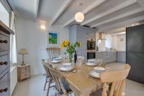 A dining area with a wooden table set for four, surrounded by chairs. There is a vase with flowers on the table, and a painting on the wall above it. - 3 Bed in Gunwalloe (oc-p00858) (Gunwalloe)