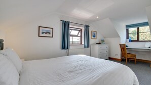 A bedroom with a large bed, a desk with a chair, a radiator, and a window with curtains. - 3 Bed in Gunwalloe (oc-p00858) (Gunwalloe)
