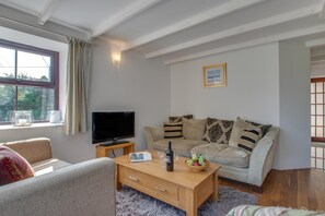 A living room with a sofa, a coffee table with a bottle and glasses, a TV on a stand, and a window with curtains. - 3 Bed in Gunwalloe (oc-p00858) (Gunwalloe)