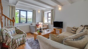 A cozy living room with a sofa set, a coffee table with a fruit bowl, a television on a stand, and a window with curtains. - 3 Bed in Gunwalloe (oc-p00858) (Gunwalloe)