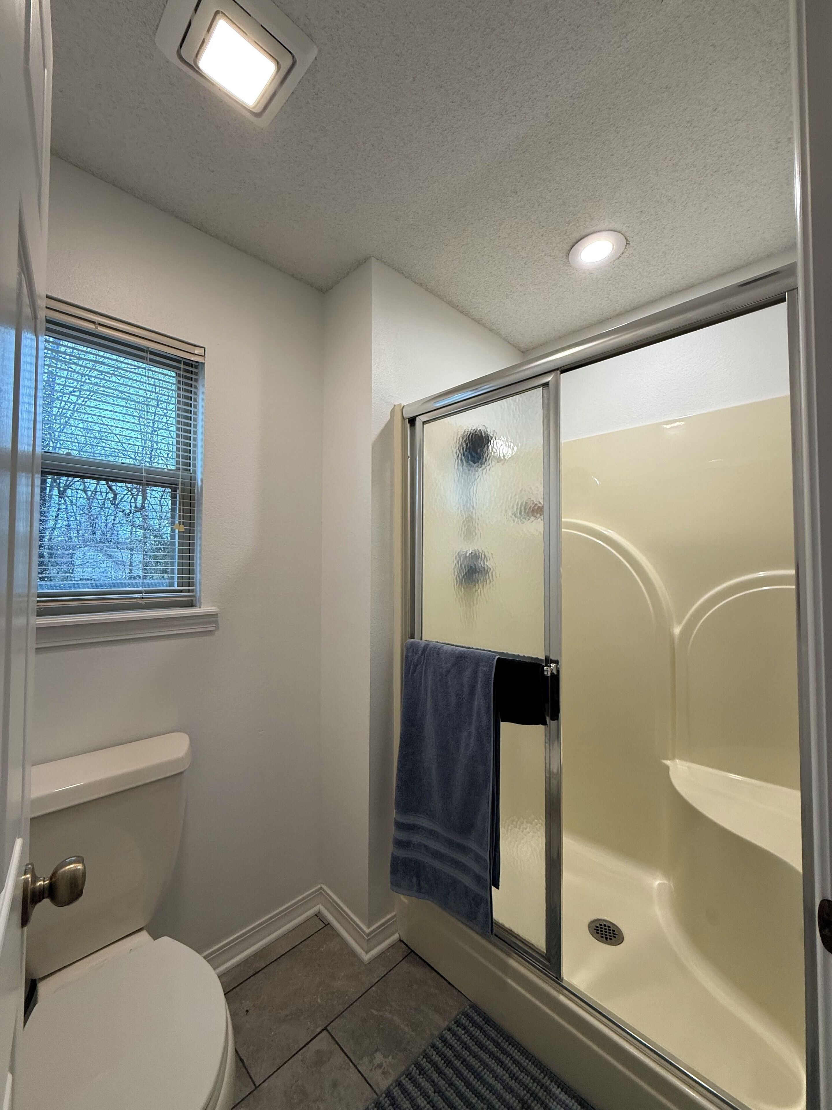 A bathroom with a toilet, a glass-enclosed shower, and a window with blinds.