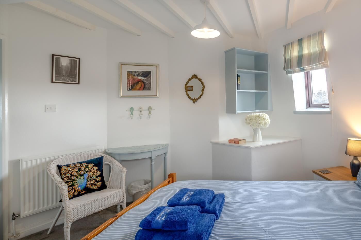 Ein Schlafzimmer mit einem Bett, einem Sessel, einem kleinen Tisch, einem Regal mit einer Vase und einem Fenster mit Vorhängen.