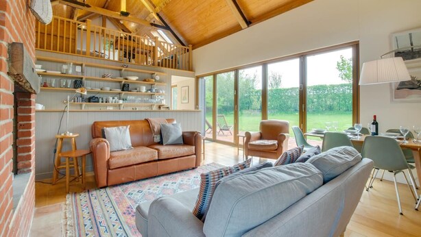A spacious living room with a high ceiling, a wooden staircase, and a large window offering a view of the outdoors. - 3 Bed in Debenham (oc-thefol) (Debenham)
