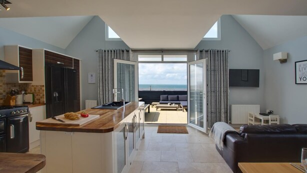 Eine moderne Küche mit Kochinsel, ein Essbereich mit Tisch und ein Wohnzimmer mit Sofa. Eine große Schiebeglasdoor führt zu einem Balkon mit Meerblick.
