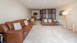 A living room with a brown leather sofa, a green armchair, and a brown sofa with patterned cushions. There is a wooden table with a lamp, a radiator, and a painting on the wall. - 4 Bed in Saundersfoot (oc-pw9197) (Saundersfoot)
