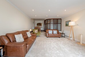 Ein Wohnzimmer mit braunem Ledersofa, einem grünen Sessel und einem weiteren braunen Sofa mit gemusterten Kissen. Ein Holztisch mit Lampe, Heizkörper und einem Bild an der Wand sind ebenfalls zu sehen.